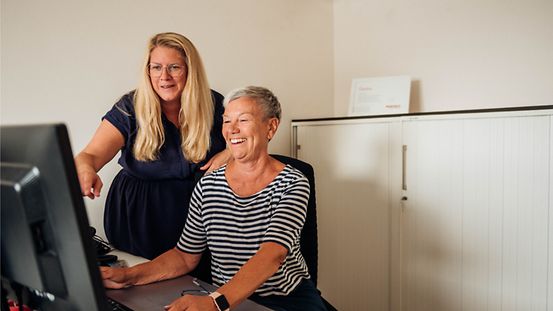 Linda Marx sitzt an einem Arbeitsplatz mit Computer, links neben ihr steht ihre Chefin Vanessa Weber.