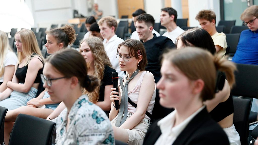 Auf dem Bild 15 junge Personen zu sehen, welche in vier Reihen hintereinander sitzen. Eine Person hält ein Mikrophon und spricht.