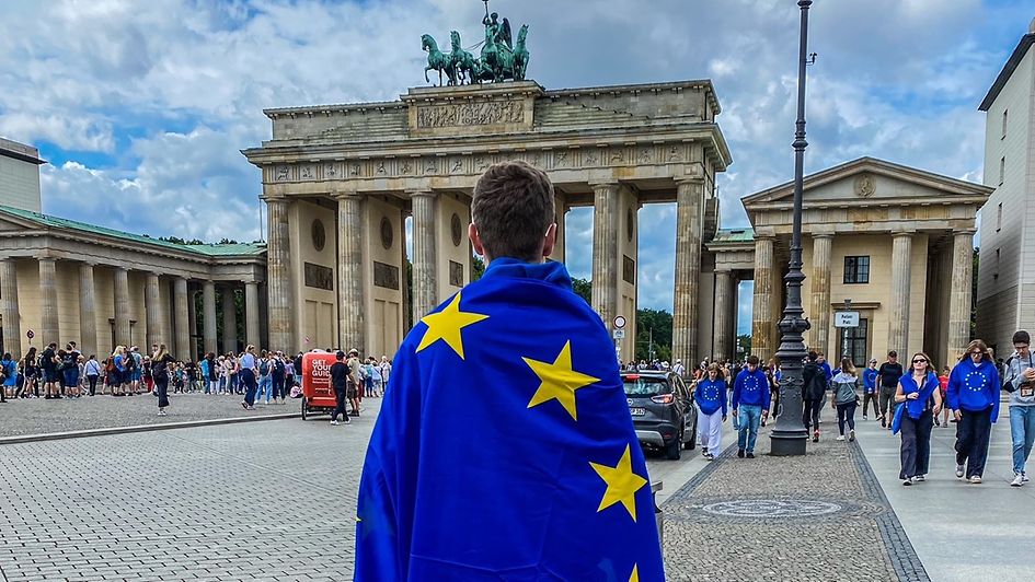 ein junger Mann steht in einer EU-Flagge umhüllt vor dem Brandenburger Tor
