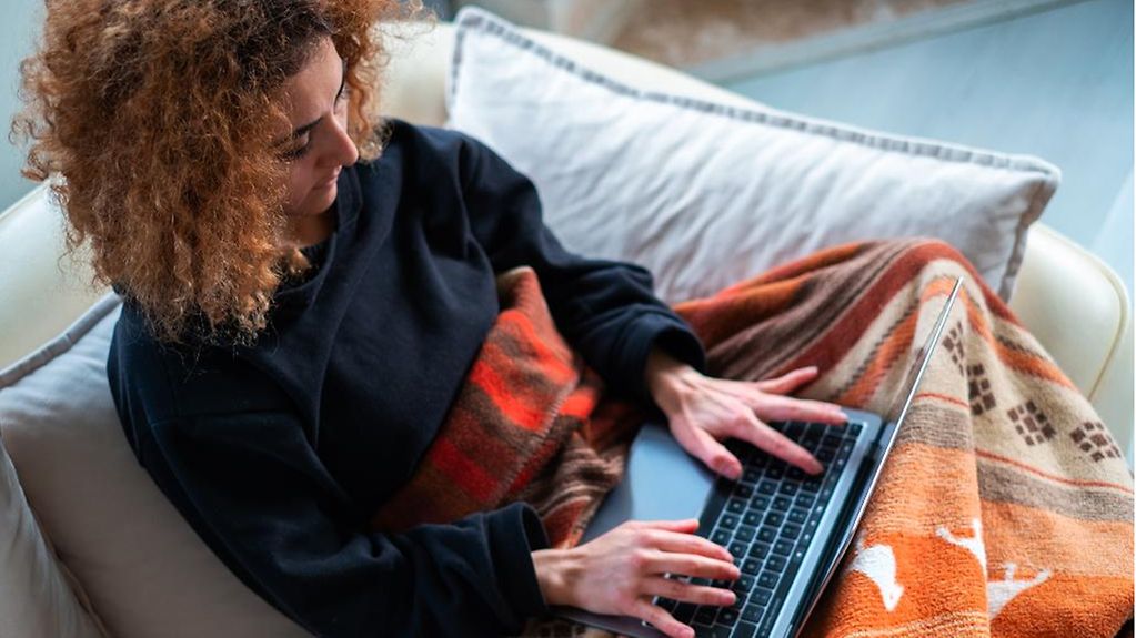 Eine junge Frau sitzt eingewickelt in einer Decke auf der Couch. Auf ihrem Schoss ein Laptop.