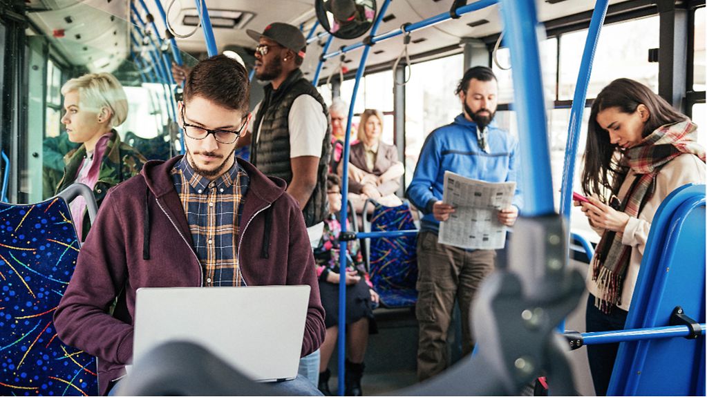 In einem Bus blicken verschiedene Fahrgäste auf einen Laptopbildschirm, in eine Zeitung und auf ein Handy.