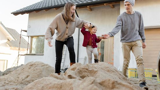 Eltern spielen mit ihrem Kind auf einem Sandhügel vor ihrem neuen Eigenheim.