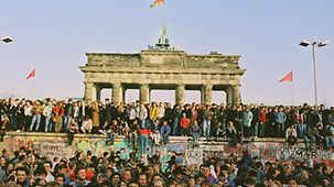 People from West and East Berlin stand on and beside the Wall at the Brandenburg Gate.