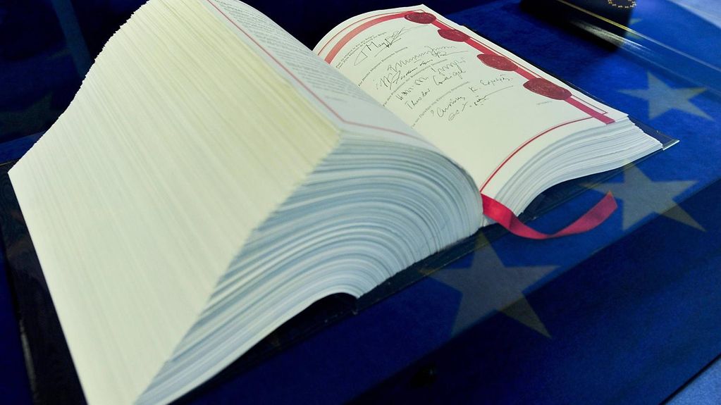 Ein dickes Buch liegt geöffnet auf einem blauen Untergrund mit goldenen Sternen. Im Buch steht der Vertrag von Maastricht.