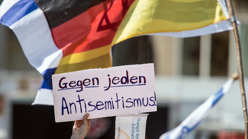 Eine Hand hält ein Schild aus Pappe hoch. Auf dem Schild steht: Gegen jeden Antisemitismus. Dahinter sieht man die Flagge von Deutschland über der Flagge von Israel.