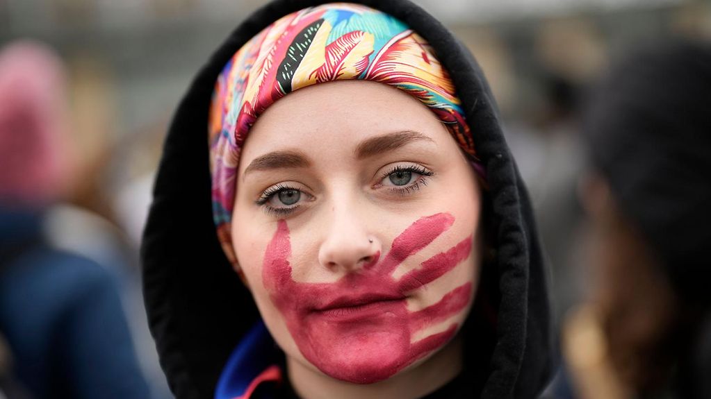 Eine junge Frau blickt in die Kamera, auf ihrem Gesicht hat sich einen roten Handabdruck gemalt.