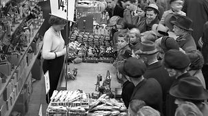 In einem Spielzeugladen oder in der Spielzeugabteilung eines Kaufhauses drängen sich Kinder und Erwachsene an einem Verkaufsstand. Eine Verkäuferin scheint etwas zu zeigen oder vorzuführen.