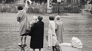 Vier Frauen stehen in der Bernauer Straße vor der Berliner Mauer. Eine steht auf einem Klapphocker, winkt mit einem Tuch. Eine andere trägt ein Baby, eine weitere hat einen Kinderwagen in der Hand. 