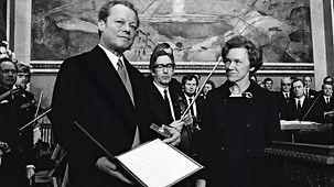 Willy Brandt hält den Friedensnobelpreis und die Urkunde in den Händen. Neben ihm steht die Vorsitzende des Nobelpreis-Komitees Aase Lionäs. Im Hintergrund stehen Musiker mit ihren Instrumenten. 
