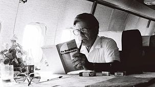 Helmut Schmidt sitzt in einem Flugzeug an einem Tisch und liest in einem Buch.