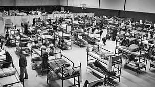Viele Menschen in einer Turnhalle, die mit vielen Tischen und Stühlen zu einer Kantine umfunktioniert worden ist.