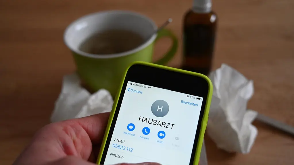 Eine Hand hält ein Handy in der Hand. Auf dem Bildschirm steht „Hausarzt“. Im Hintergrund steht eine grüne Tasse Tee auf einem Tisch. Daneben sind Papiertaschentücher und eine Flasche Medizin. 
