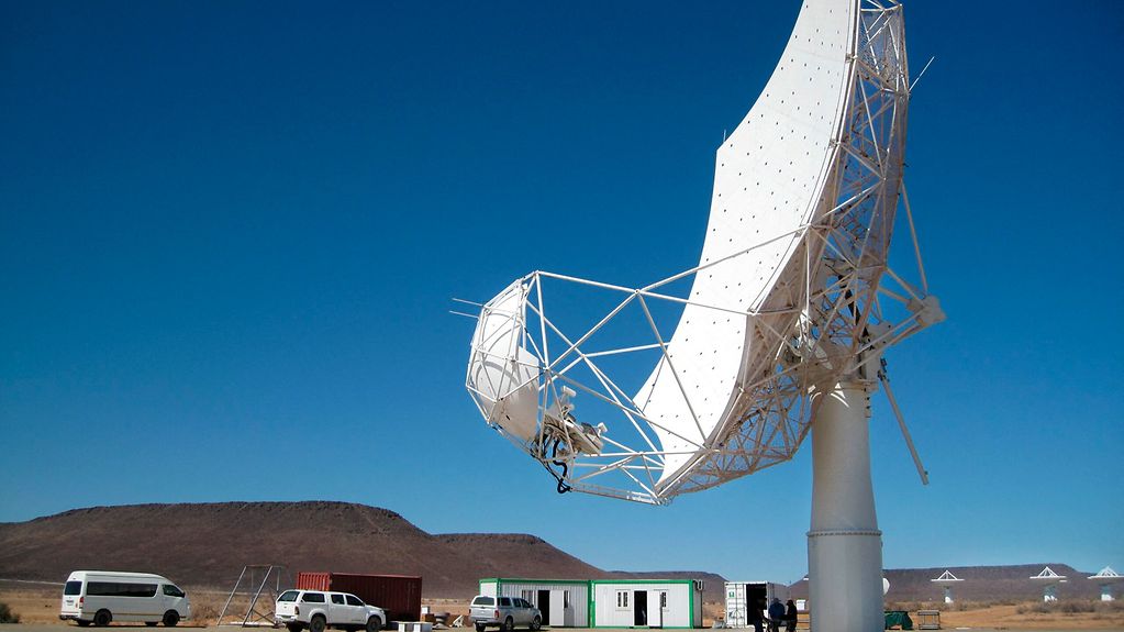 Bild zeit ein großes Radioteleskop in einer Wüstenlandschaft im Hintergrund. Einige Fahrzeuge, Baucontainer und kleinere Teleskope sind ebenfalls zu sehen.