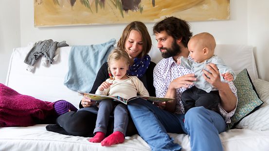 Eine vierköpfige Familie mit zwei Kleinkindern sitzt auf dem Sofa und schaut sich ein Buch an.