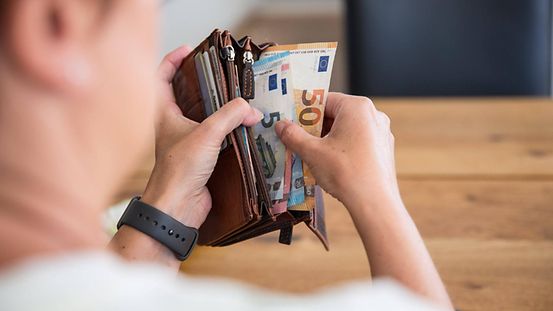 Das Foto ist über die Schulter eine Person fotografiert, deren Silhouette noch erkennbar ist und die einen geöffneten Geldbeutel mit mehreren Euro-Scheinen in den Händen hält.