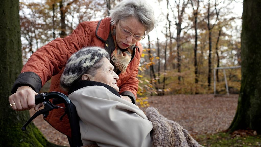 Frau mit ihrer 94-jährigen Mutter im Rollstuhl in einem Wald.