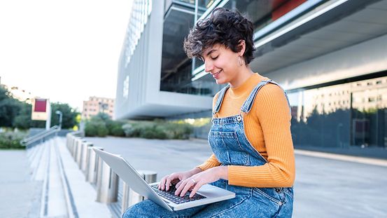 Eine junge Frau sitzt auf einem metall Poller vor einem Gebäude und tippt auf eine Laptop.