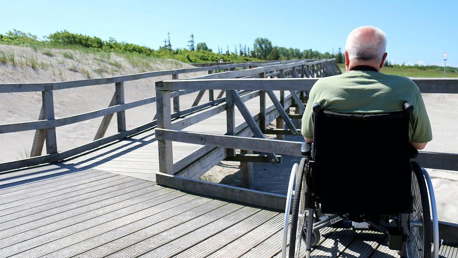 Ein Mann sitzt im Rollstuhl auf einem Holzsteg am Strand.