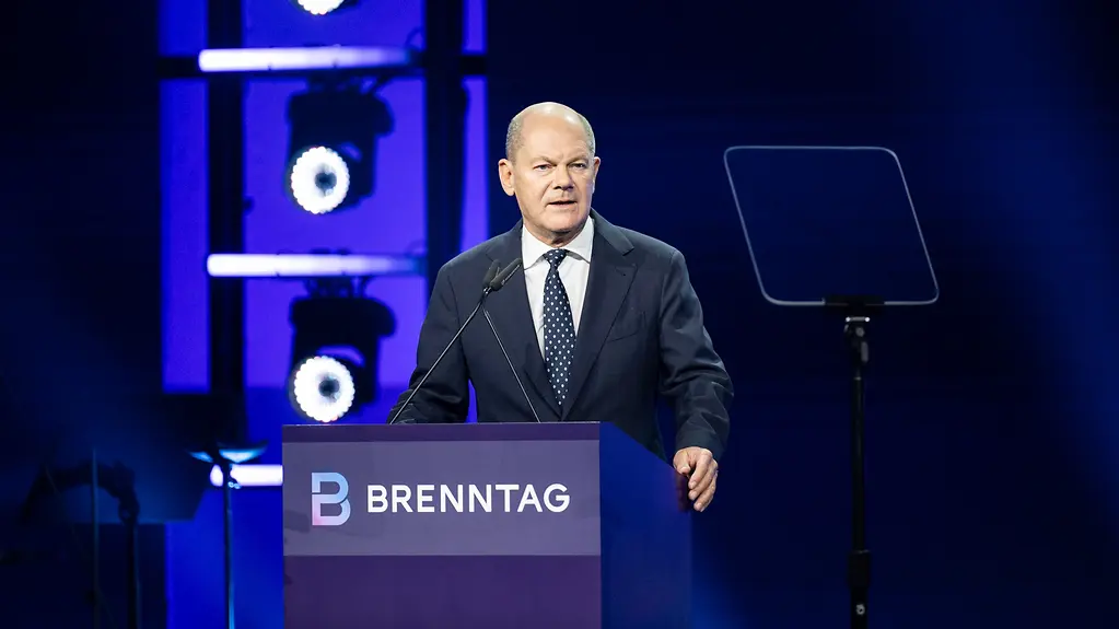 Bundeskanzler Scholz spricht auf einer dunkelblau beleuteten Bühne an einem Rednerpult mit der Aufschrift "Brenntag". Rechts von ihm ein Telepromter.