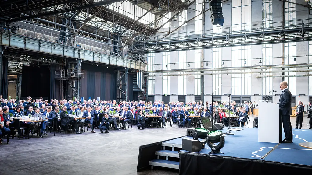 Blick von der Seite in eine Halle. Auf der linken Seite sind viele Tische aufgestellt, die voll besetzt sind. Auf der rechten Seite spricht der Kanzler auf einer Bühne an einem Rednerpult. 