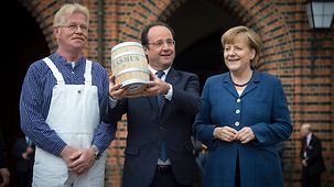 Der französische Präsident François Hollande mit einem Fass Heringe mit Fischer Rasmus und Bundeskanzlerin Angela Merkel.