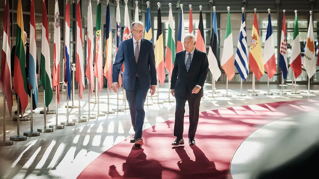 Bundeskanzler Merz und EU-Ratspäsident Costa gehen über einen roten Teppich vor den Flaggen der EU-Staaten.