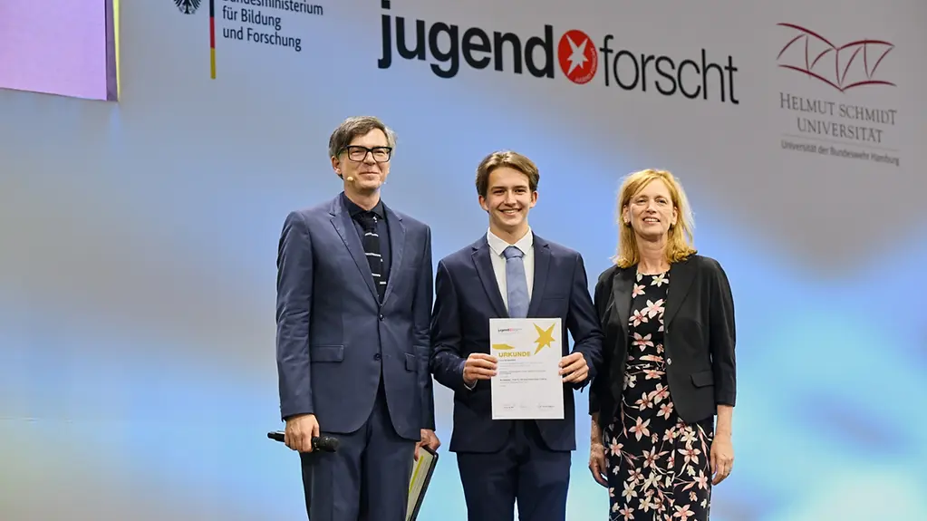 Preisträger Louis Schwarzlose mit Bundesbildungsministerin Karin Prien und Moderator Ralph Caspers auf der Bühne