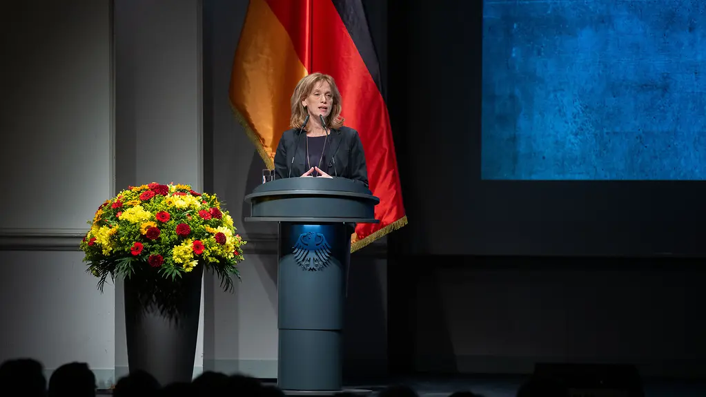 Bundesfrauenministerin Karin Prien spricht an einem Rednerpult im Hintergrund eine Deutschlandflagge. Rechts Blumenschmuck. 