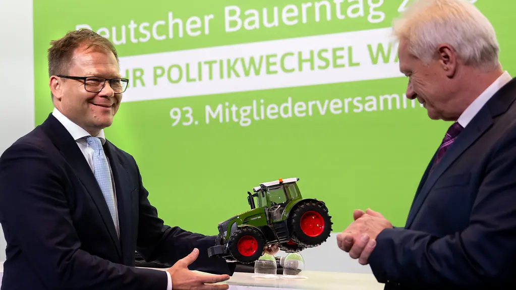Carsten Schneider übergibt und Alois Rainer einen Spielzeugtraktor vor grünem Hintergrund.