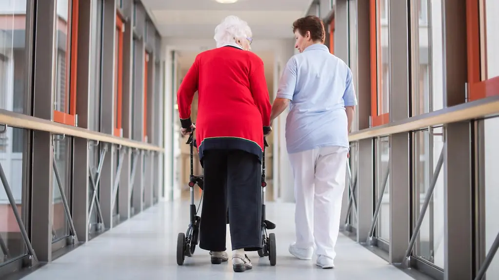 Auf dem Foto zu sehen ist eine Pflegerin, die mit einer Bewohnerin des Pflegeheims und einem Rollator über den Flur geht.