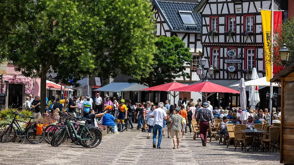 Auf dem Foto zu sehen sind Menschen in einer Stadt unter Schirmen in Gastronomieeinrichtungen sitzend.