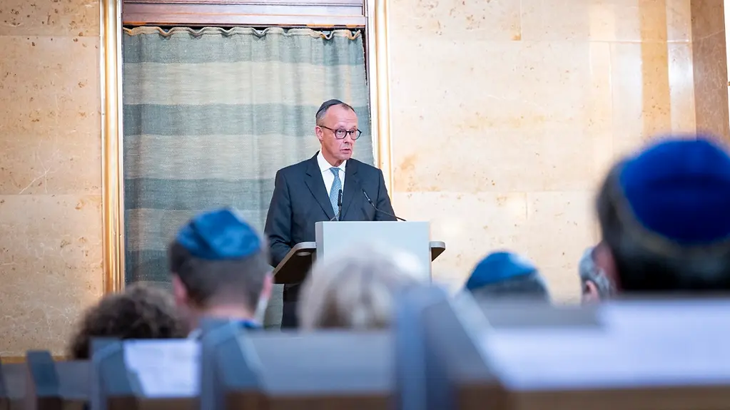 Le chancelier fédéral Merz coiffé d’une kippa derrière son pupitre dans la synagogue. En arrière-plan, une fenêtre avec une inscription en hébreu.