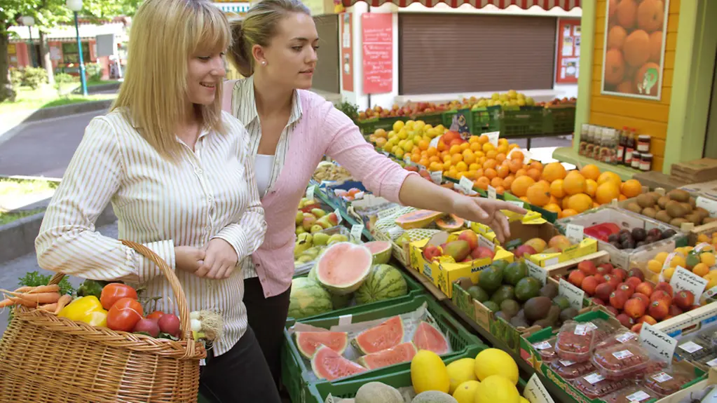 Zwei junge Frauen beim Einkaufen an einem Obststand im Freien.