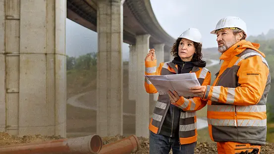 Eine Frau und ein Mann mit Helm und Warnkleidung stehen konzentriert mit einem Plan in den Händen neben einer großen Brücke. 