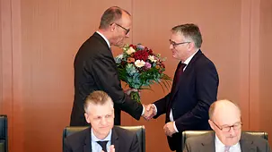 Das Foto zeigt Bundeskanzler Friedrich Merz, der Stefan Kornelius, Sprecher der Bundesregierung, einen Blumenstrauß überreicht.