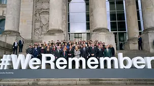 Das Foto zeigt Bundeskanzler Friedrich Merz und weitere Menschen hinter dem Claim #WeRemember anlässlich des Holocaust-Gedenktags.