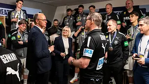 Das Foto zeigt Bundeskanzler Friedrich Merz in der Kabine der deutschen Mannschaft mit Spielern um Bundestrainer Alfred Gislason.