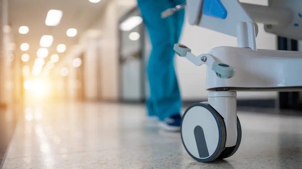 Ein Krankenhausflur mit einer Pflegekraft, die einen Wagen schiebt. Der Flur ist leer und die Sonne scheint durch die Fenster.