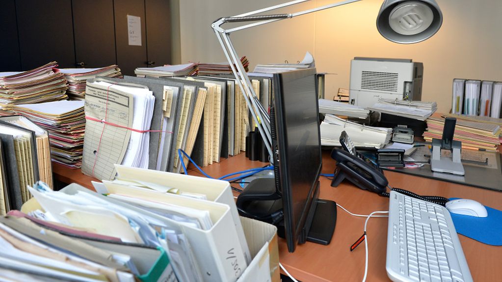 Büro mit Schreibtisch voll Akten, Aktenordner und dazwischen ein Computer