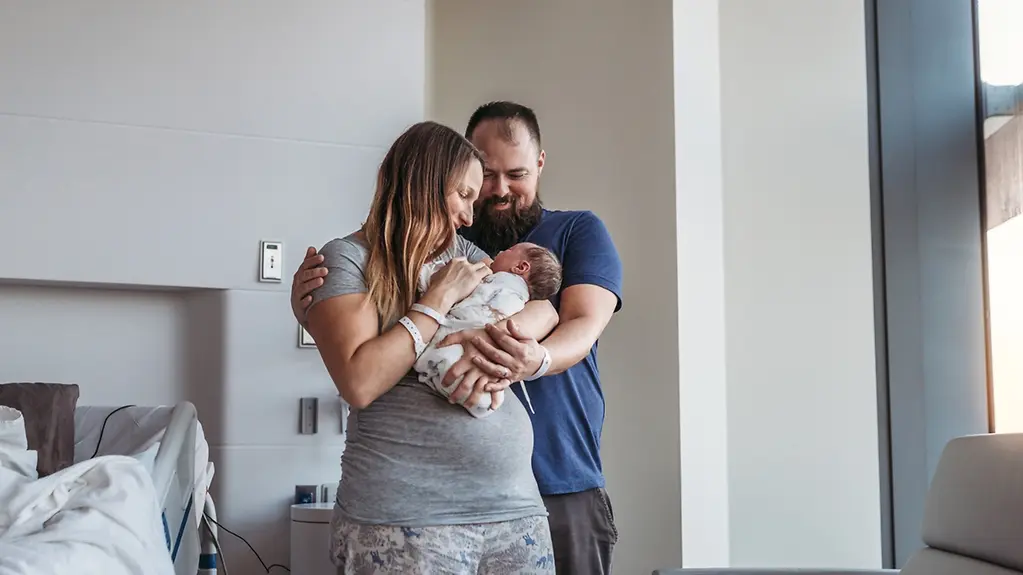 Eine junge Mutter hält ein Neugeborenes im Arm, hinter den beiden steht der Vater. 
