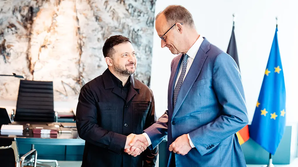 Zelenskyy and Merz shake hands and look each other in the eye with a smile.