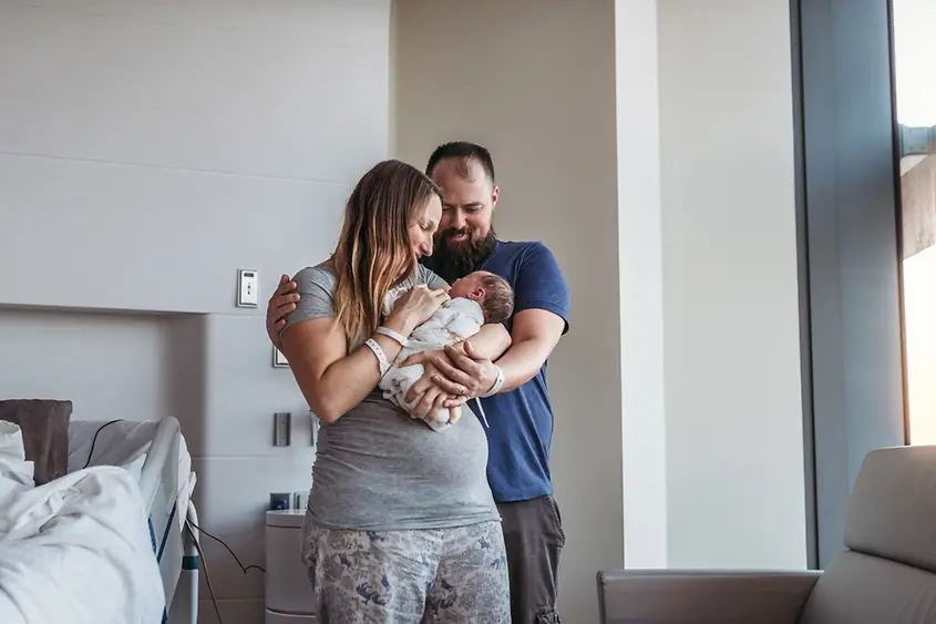 Eine junge Mutter hält ein Neugeborenes im Arm, hinter den beiden steht der Vater. 