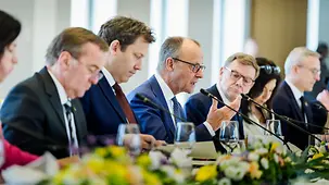 Chancellor Merz speaks into a microphone, with ministers of the Federal Government sitting next to him.