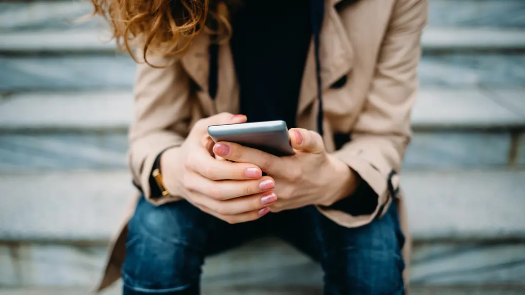 Nahaufnahme einer jungen Frau in einem beigen Trenchcoat und blauen Jeans, die auf einer grauen Steintreppe sitzt und ihr Smartphone benutzt.
