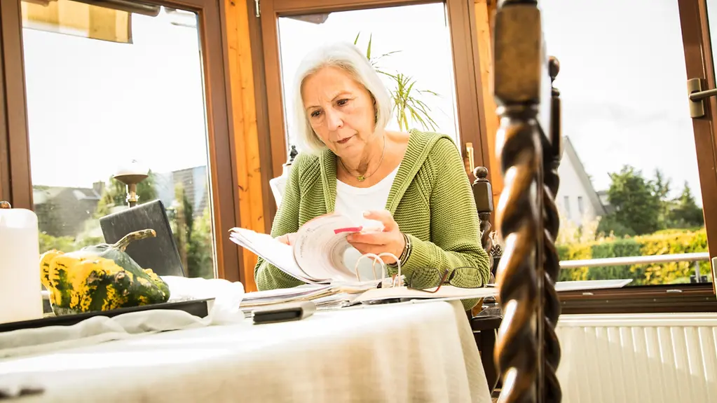  Eine ältere Frau sitzt an ihrem Schreibtisch und sortiert Belege.