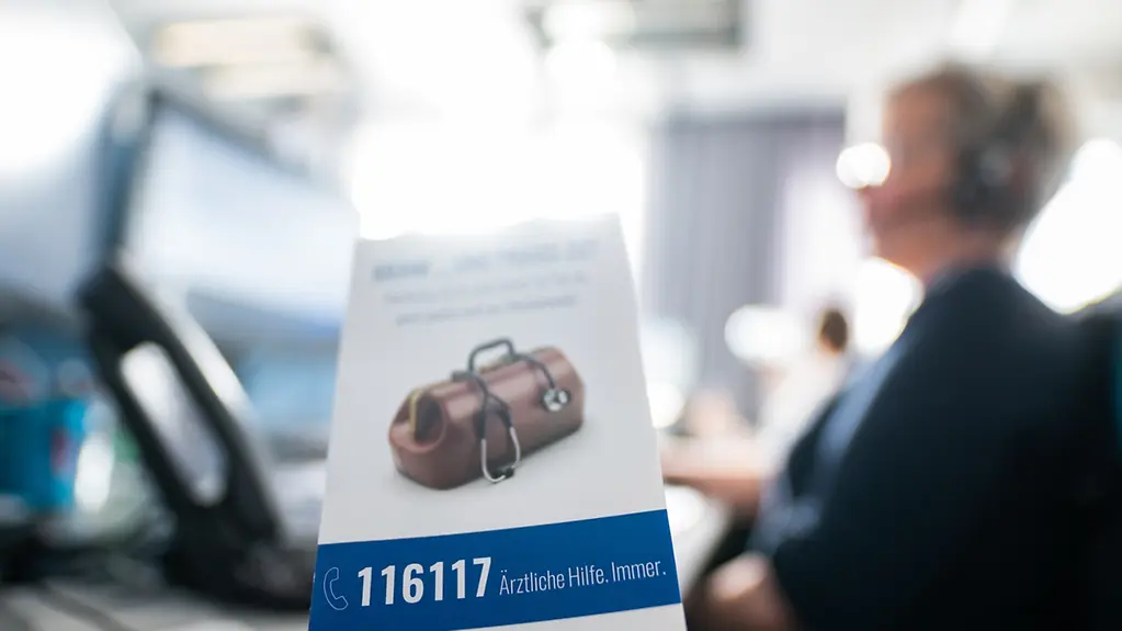 Flyer für die Rufnummer des ärztlichen Bereitschaftsdienstes «116117», im Hintergrund unscharf eine telefonierende Person in einer Telefonzentrale für den Bereitschaftsdienst der Kassenärztlichen Vereinigung Hamburg