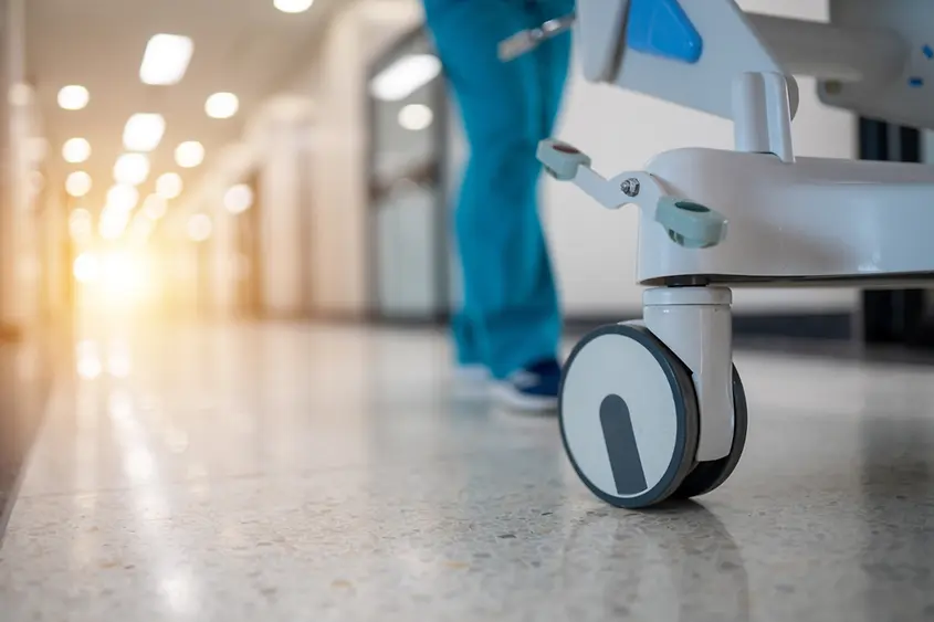 Ein Krankenhausflur mit einer Pflegekraft, die einen Wagen schiebt. Der Flur ist leer und die Sonne scheint durch die Fenster.