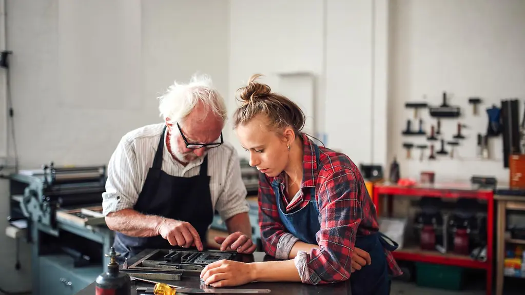Ein älterer Mitarbeiter und eine junge Mitarbeiterin stehen gemeinsam an einem Arbeitstisch.