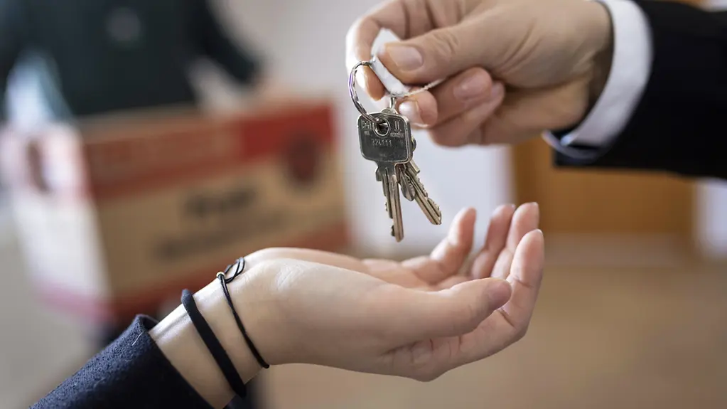 Eine Person übergibt einer anderen Person den Schlüssel für eine Wohnung, im Hintergrund sind Umzugskartons zu sehen.