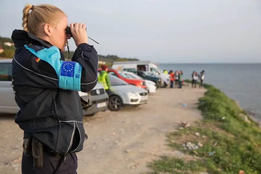 Eine deutsche Bundespolizistin mit Frontex-Armbinde steht an der Küste und schaut durch ein Fernglas aufs Meer.